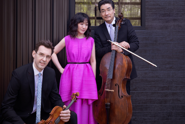Three people next to each other. A man in a suit is sitting on the left and is holding his violin. In the middle is a woman in a pink dress posing. On the right a man in a suit is posing with his cello.